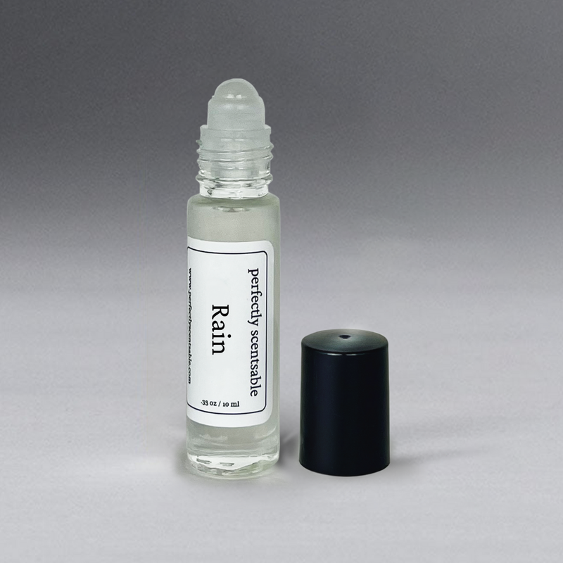 A small, transparent roller bottle labeled "Perfectly Scentsable Rain Perfume Oil - Rollerball," containing clear liquid, with a black cap placed beside it. The bottle holds 10 ml, as indicated on the label. The background is a gradient of gray shades. This gender-neutral scent by Perfectly Scentsable is perfect for any occasion.