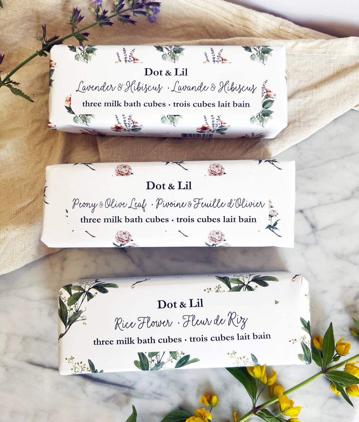 The Dot & Lil Lavender & Hibiscus Milk Bath Cubes Trio Gift Set is displayed on a marble surface with flowers and a beige cloth.