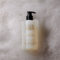 A pump bottle of Caswell-Massey Almond & Aloe Hand & Body Wash stands atop white soap suds, bubbles clinging to its label—a plant-based wash from Caswell-Massey for indulgent botanical skincare routines.