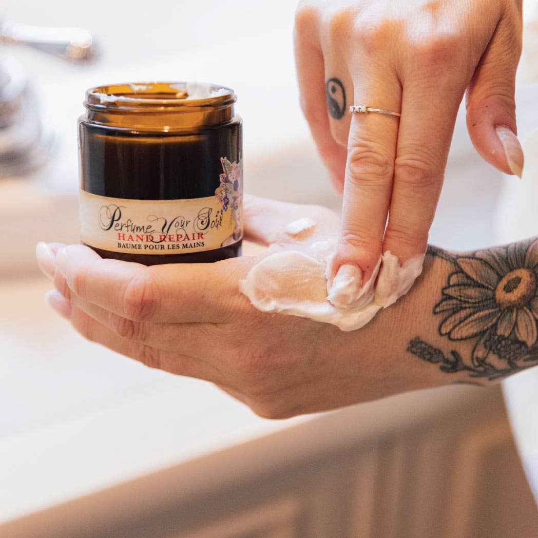 A person with a small ring and a yin-yang tattoo on their finger applies Barefoot Venus Lemon Freckle Hand Repair Balm from a brown jar onto the back of their hand.