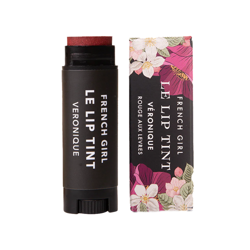 A tube of FRENCH GIRL Tinted Lip Balm in Veronique, a reddish shade, is placed beside its floral-patterned box. Both the tube and packaging feature the FRENCH GIRL product name in white lettering.