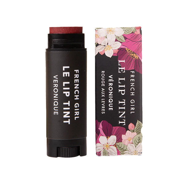A tube of FRENCH GIRL Tinted Lip Balm in Veronique, a reddish shade, is placed beside its floral-patterned box. Both the tube and packaging feature the FRENCH GIRL product name in white lettering.