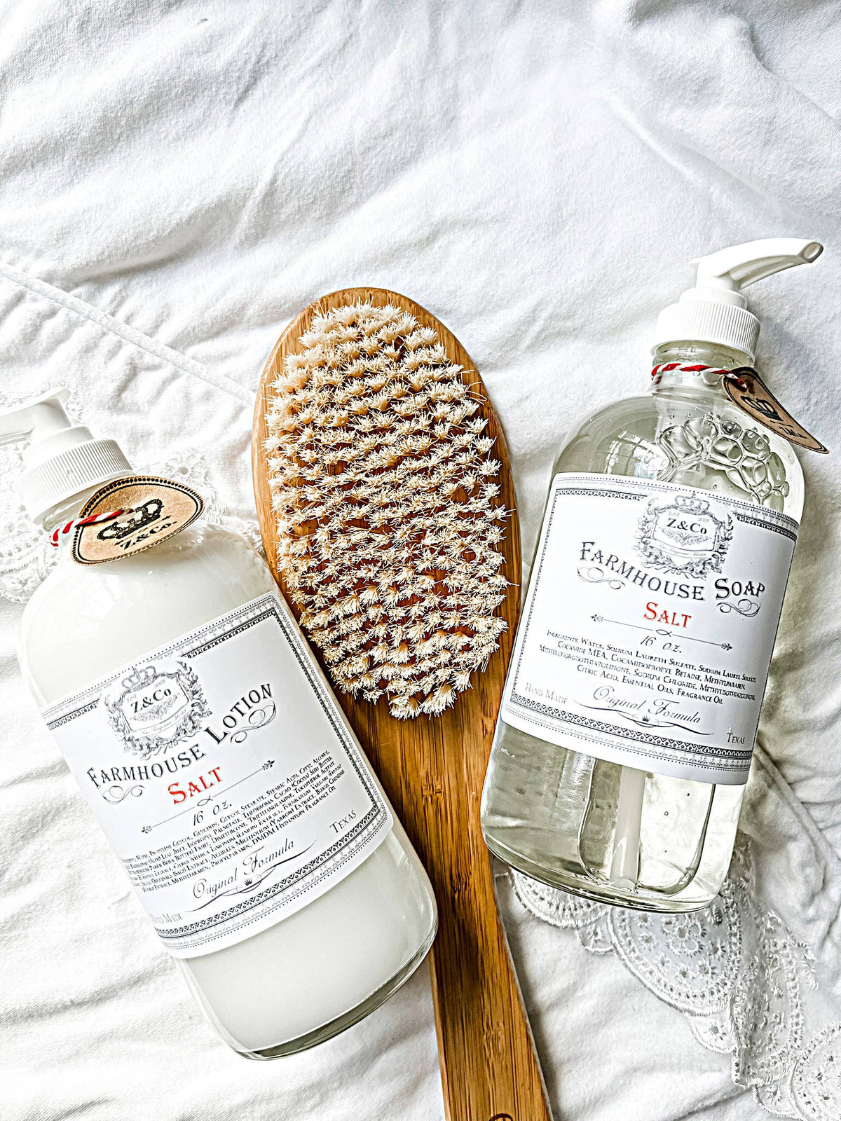 A flat lay image showcases a bottle of Z&Co. Hey There Pumpkin Farmhouse Fall Soap and a bottle of farmhouse lotion, both labeled "salt," placed on white lace fabric. A wooden-handled body brush with bristles is positioned between the two bottles, evoking cozy vibes of pumpkin pie and creamy vanilla.