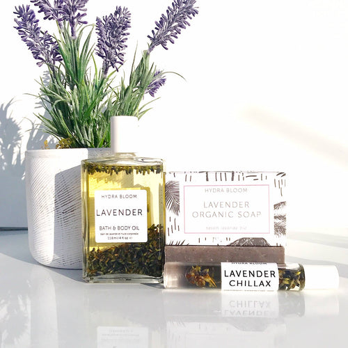 Hydra Bloom Beauty Lavender-themed bath products including organic hand soap, soap bars, and a vase with artificial lavender flowers on a white surface with soft light.