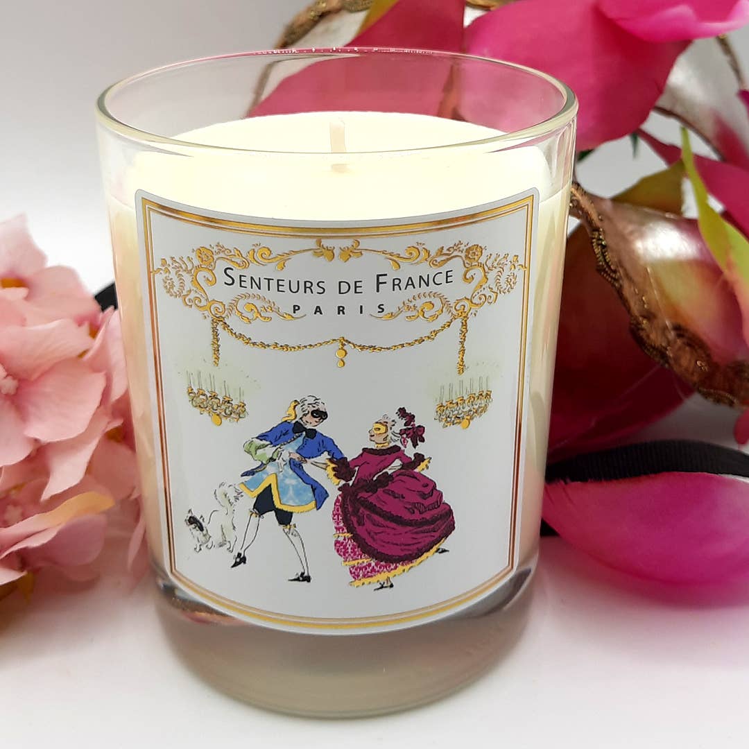 The Senteurs De France Violet Candle Royal Masquerade Ball features an 18th-century dancing couple on its jar, is made from vegetable wax, infused with Grasse fragrance, and reads "Senteurs de France Paris," with pink flowers arranged beside it.
