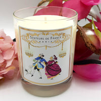 The Senteurs De France Violet Candle Royal Masquerade Ball features an 18th-century dancing couple on its jar, is made from vegetable wax, infused with Grasse fragrance, and reads "Senteurs de France Paris," with pink flowers arranged beside it.
