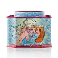 A rectangular tin container with a vintage-style illustration on the front. The illustration depicts a woman in a blue dress reclining on a pillow. Labeled "Barefoot Venus," with the scent "Coconut Kiss Bath Soak," crafted with paraben and sulfate-free, plant-based ingredients, it features blue and pink accents.