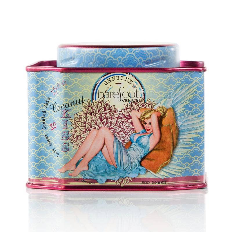 A rectangular tin container with a vintage-style illustration on the front. The illustration depicts a woman in a blue dress reclining on a pillow. Labeled "Barefoot Venus," with the scent "Coconut Kiss Bath Soak," crafted with paraben and sulfate-free, plant-based ingredients, it features blue and pink accents.