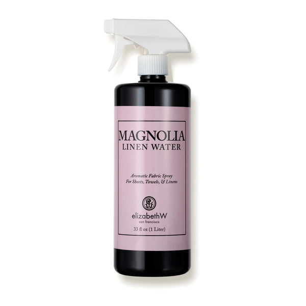 A black spray bottle of "elizabeth W Signature Magnolia Linen Water" by elizabeth W, with a pink label showing usage instructions for sheets, towels, and linens, isolated on a white background.
