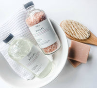 A white bowl contains a folded white towel, a glass bottle of pink Himalayan bath salts, a bottle of Z&Co. Ritual Market Collection Bubble Bath, a wooden body brush with bristles, and a bar of pink soap. The photo captures an essential oils-enhanced spa-like bath setup on a white surface.