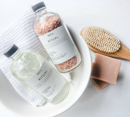 A white bowl contains a folded white towel, a glass bottle of pink Himalayan bath salts, a bottle of Z&Co. Ritual Market Collection Bubble Bath, a wooden body brush with bristles, and a bar of pink soap. The photo captures an essential oils-enhanced spa-like bath setup on a white surface.