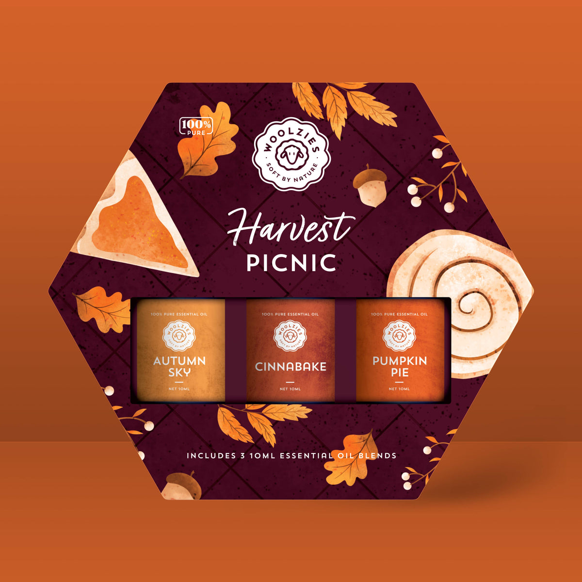 The Woolzies Harvest Picnic Essential Oil Collection is a hexagonal gift box featuring three autumn oils—Autumn Sky, Cinnabake, and Pumpkin Pie—set against a fall-themed background with leaves, pie, and a cinnamon roll.