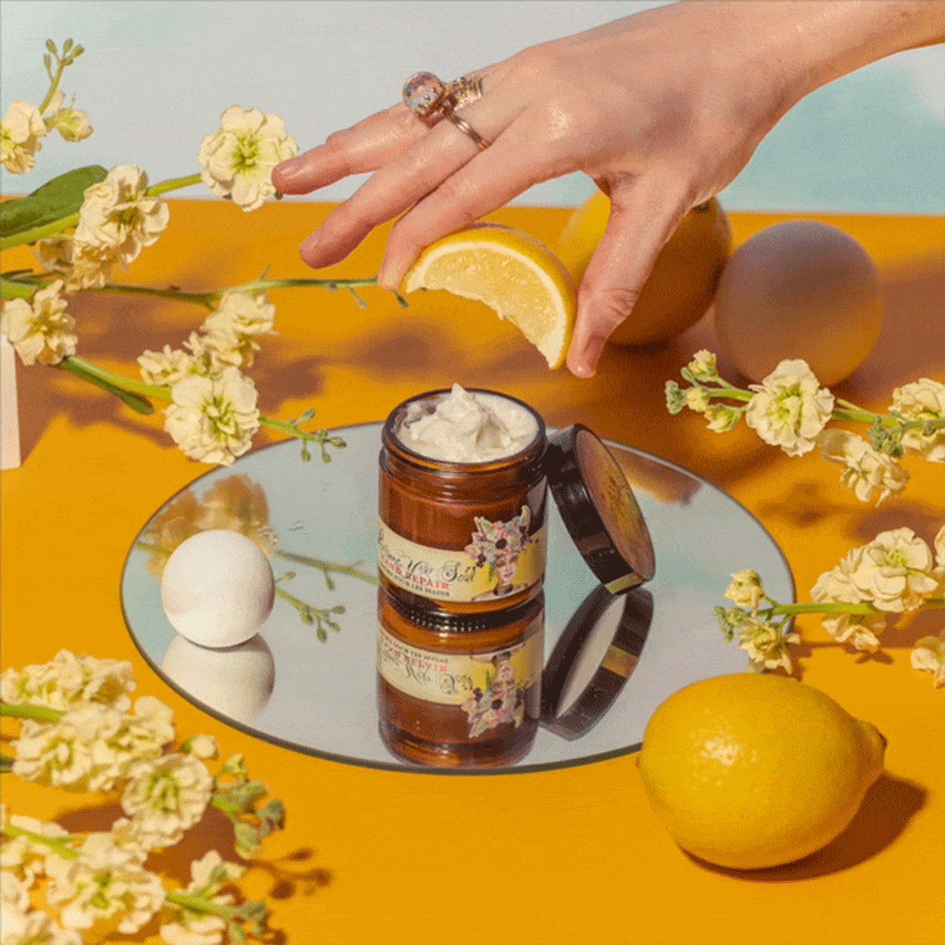 A hand with a lemon wedge hovers over an open jar of Barefoot Venus Lemon Freckle Hand Repair Balm, displayed on a round mirror and surrounded by whole lemons and yellow flowers on a vibrant yellow surface.