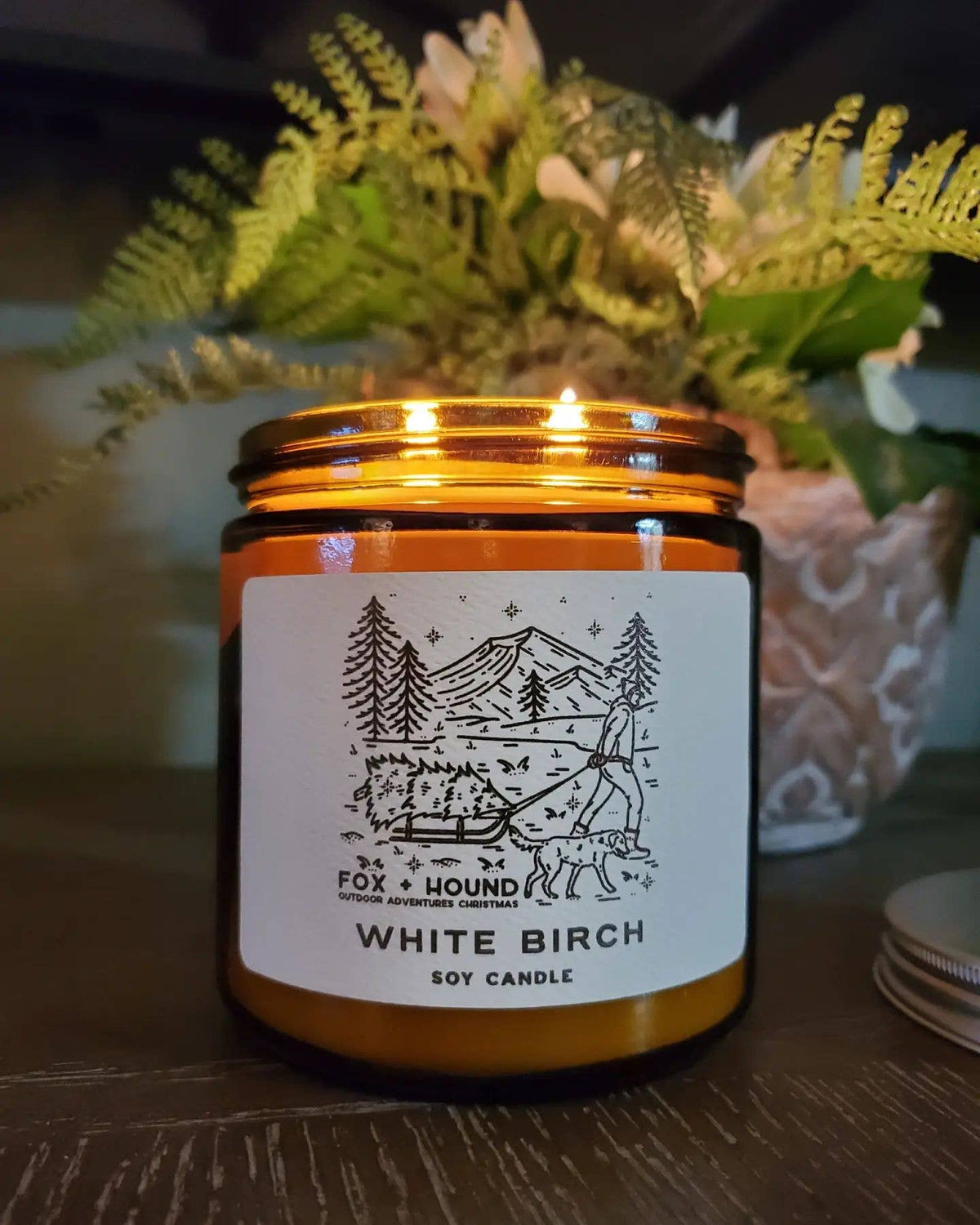 A Fox + Hound Odor Eliminator Soy Candle - White Birch sits on a table, its mountain and forest label evoking the outdoors. This candle by Fox + Hound offers a fresh scent, with a plant and coasters in the background.