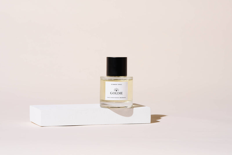 A bottle of Goldie Dog Fragrance - Summer Tonic with a black cap rests on a white platform against a light beige background, showcasing the refined style of Goldie's luxury dog grooming products.