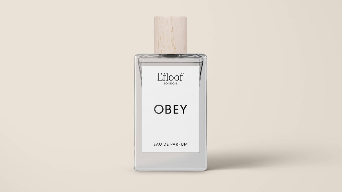 A clear glass perfume bottle with a wooden cap is displayed against a beige background. The label reads "L'floof" at the top, "OBEY" in the center, and "Eau de Parfum" at the bottom. The dog perfume spray by L'floof boasts a fruity floral scent.