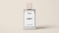 A clear glass perfume bottle with a wooden cap is displayed against a beige background. The label reads "L'floof" at the top, "OBEY" in the center, and "Eau de Parfum" at the bottom. The dog perfume spray by L'floof boasts a fruity floral scent.