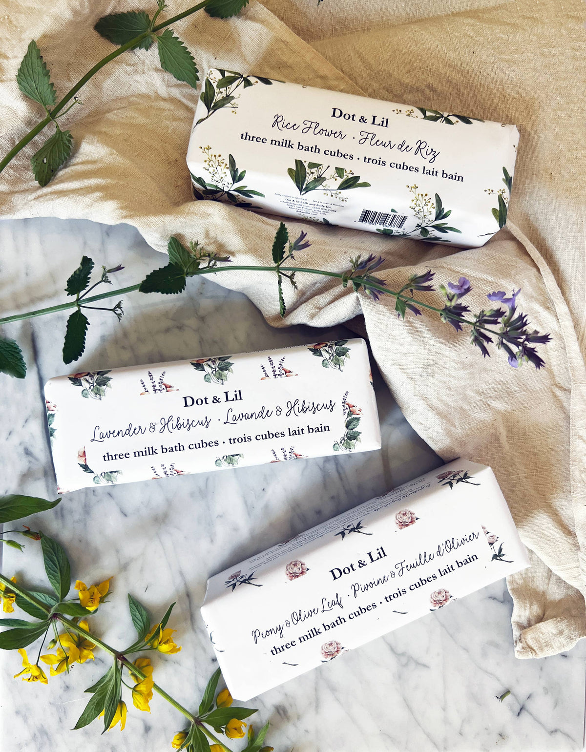 The Dot & Lil Lavender & Hibiscus Milk Bath Cubes Trio Gift Set rests on marble with a cream cloth and wildflowers. Each package features delicate floral illustrations and highlights its unique scent.