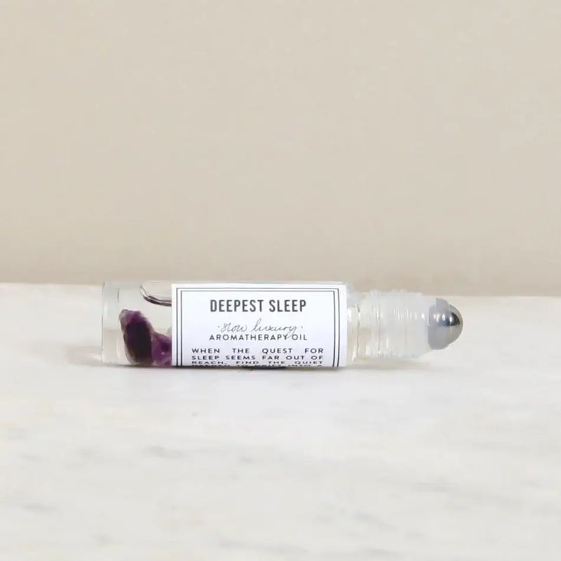 The Norfolk Natural Living Deepest Sleep Pulse Point Oil (10ml) rests on a white surface, featuring clear liquid and amethyst gemstones to support restful sleep.