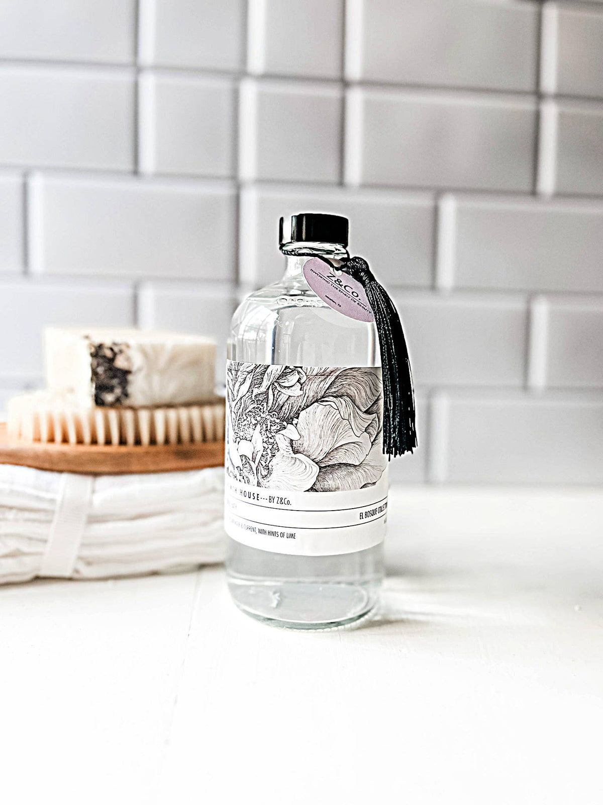 A clear bottle of liquid from the Z&Co. French House El Bosque French House Bubble Bath, adorned with a black tassel and a pink tag, sits on a white surface against a tiled background. Nearby, an all-natural fragrance soap wrapped in white paper rests beside a wooden brush with light bristles. Both items feature black and white botanical illustrations.