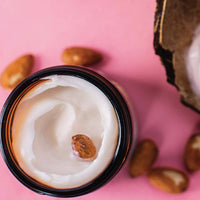 A close-up of an open jar of Barefoot Venus Coconut Kiss Instant Hand Repair Balm on a pink background with an almond placed on top, featuring the rich texture of Shea Butter. Several almonds are scattered around the jar, and part of a broken almond shell is visible in the upper right corner of the image.