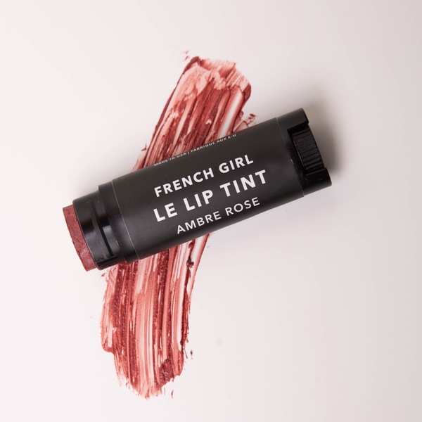A tube of FRENCH GIRL Tinted Lip Balm in Ambre Rose rests on a white surface, with a swatch of the hydrating, organic balm’s rose-tinted color displayed beneath it.