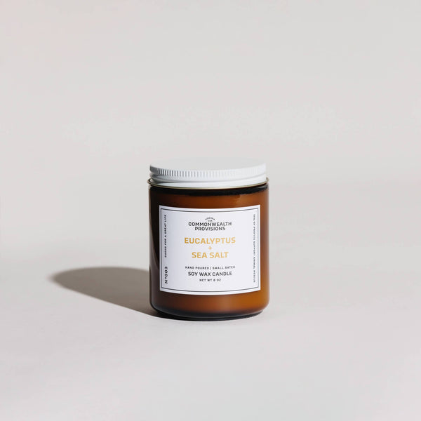 A brown glass jar candle with a white lid labeled "Eucalyptus + Sea Salt" by Commonwealth Provisions rests on a light surface, casting a soft shadow. This soy candle features phthalate-free fragrance oils for artisanal ambiance.