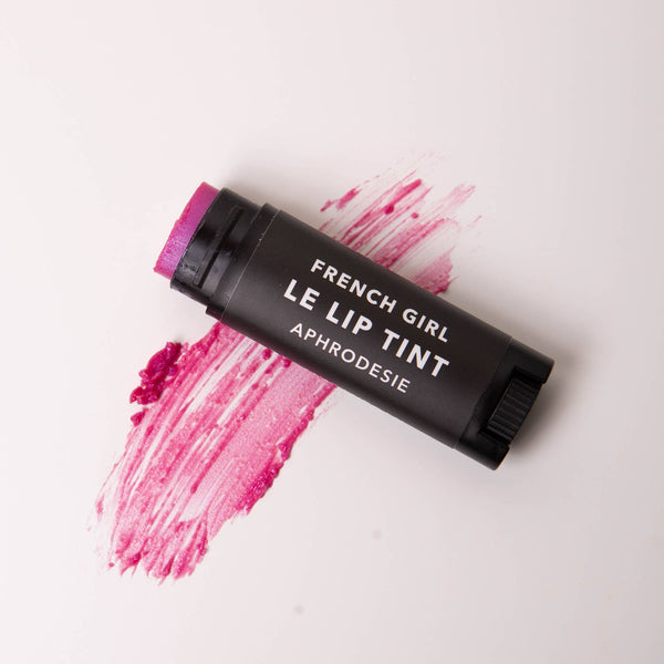 A black tube of FRENCH GIRL Tinted Lip Balm in Aphrodesie rests on a white surface, showcasing a swipe of its hydrating pink tint below.