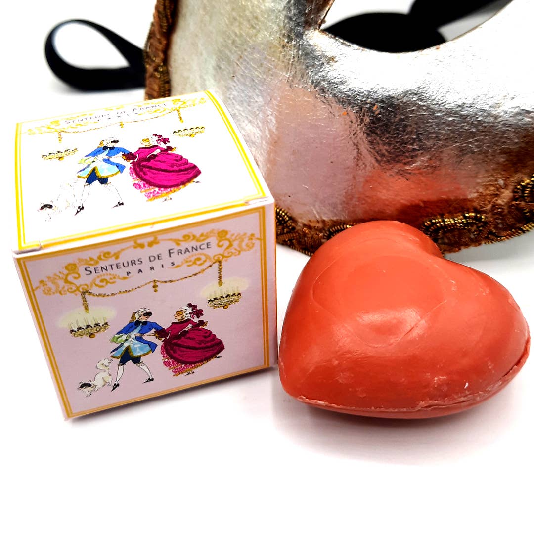The Senteurs De France Red Fruit Heart Soap sits beside its decorative box featuring a dancing couple and "Senteurs de France," evoking French heritage, while a gold-trimmed masquerade mask lies in the background.
