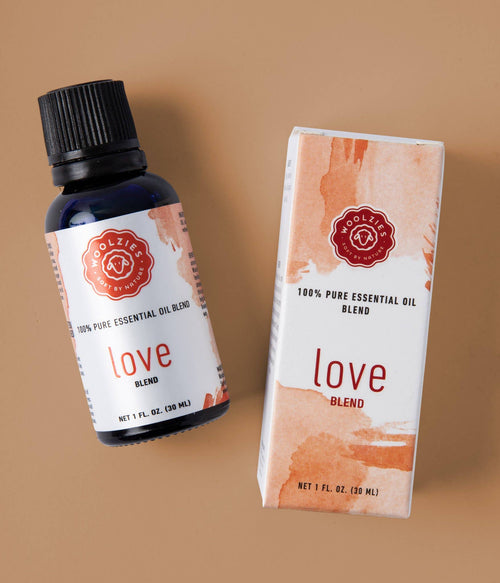 A bottle and box of Woolzies Love Blend Essential Oil, featuring a pink and white label with a red stamp logo, are displayed on a beige background.