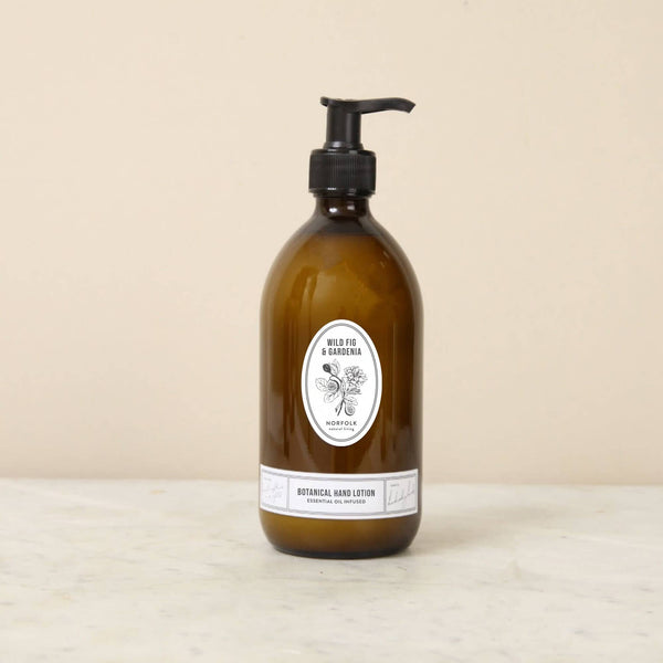 A brown glass pump bottle of Norfolk Natural Living Wild Fig & Gardenia Gardener's Hand Lotion 500ml rests on a light stone surface against a beige background.