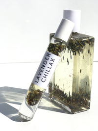 A clear glass bottle containing Hydra Bloom Beauty Lavender Chillax Organic Essential Perfume oil with lavender blossoms, labeled "lavender chillax," stands against a white background, with sunlight casting its shadow.