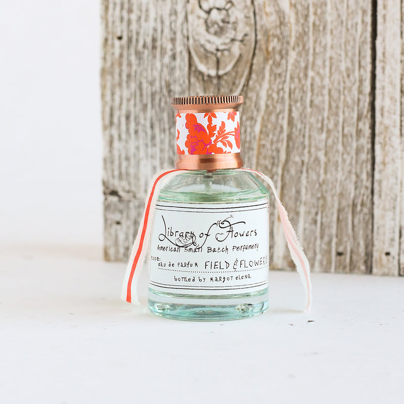 A small, clear glass bottle of artisanal Eau de Parfum labeled "Field & Flowers" by Margot Elena, with a floral design on the cork cap, against a rustic