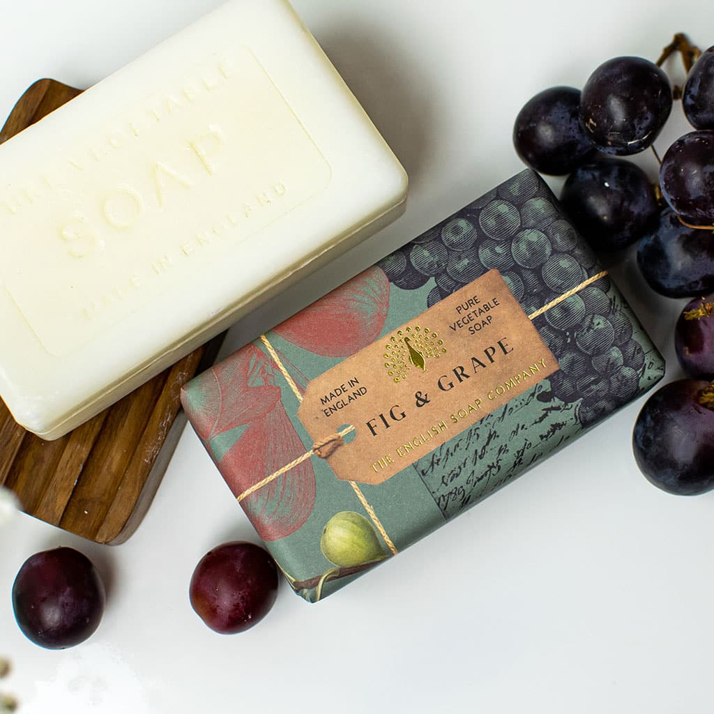 A bar of soap labeled "The English Soap Co. Anniversary Fig and Grape Soap" next to a box reading "fig & grape, pure vegetable soap," surrounded by dark and light grapes on a white surface.