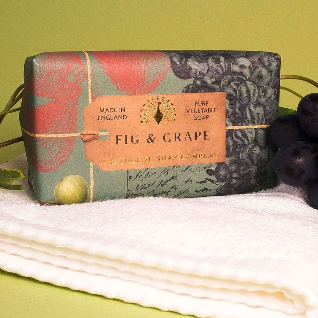 A bar of The English Soap Co. Anniversary Fig and Grape Soap, enriched with shea butter, wrapped in elegant packaging, displayed with a white towel and a cluster of grapes on a green background.
