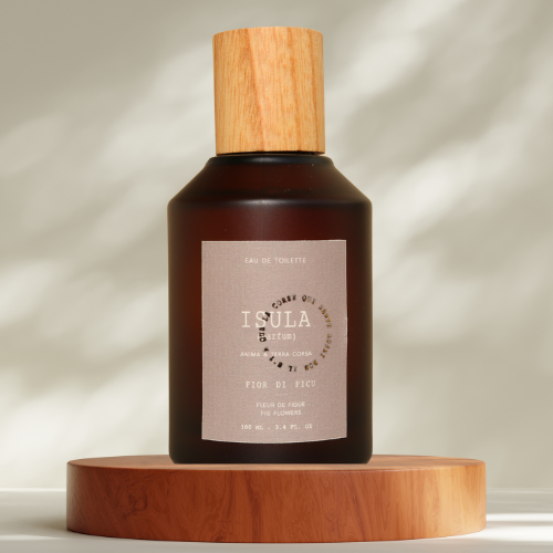 A brown ISULA parfums Fiori di Ficu - Fig Blossom eau de toilette bottle with a wooden cap rests on a round wooden platform. Its label displays the brand and scent, while soft shadows and light patterns dance in the background.