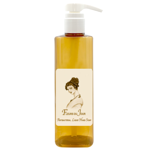 A transparent pump bottle containing amber-colored La Bouquetiere Fleurs du Jour/Marina Blue antibacterial liquid hand soap with a label featuring an illustration of a woman and the text "fleurs du jour - aromatherapy olive hair soap".