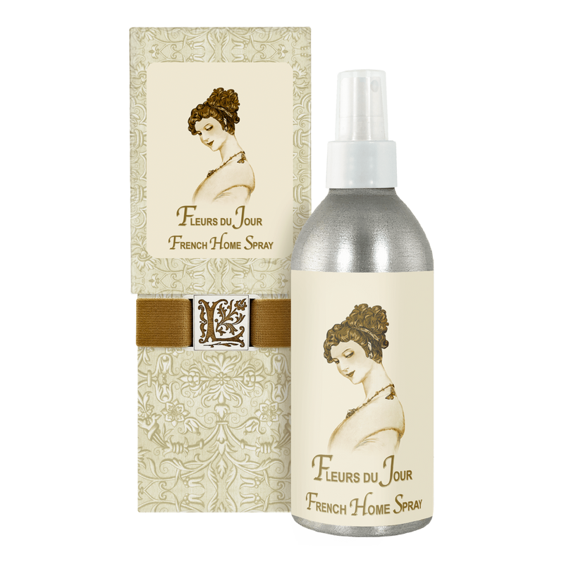 An elegant package and bottle of "La Bouquetiere Marina Bleu Home Spray" is displayed. The design features a vintage illustration of a woman with an updo hairstyle. The packaging is decorated in a floral pattern and includes a ribbon with an ornate letter "L" medallion, exuding a fresh floral fragrance.