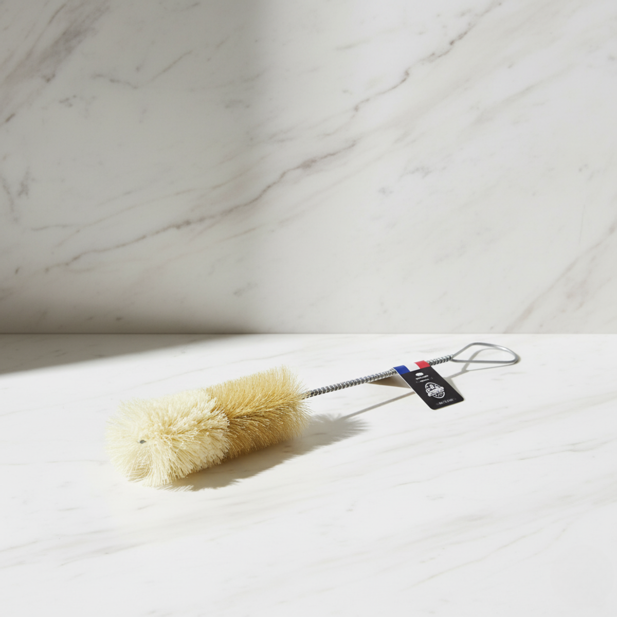 The Andrée Jardin Bistro Flute Cleaning Brush features a cream bristle head, metal handle, and black tag. Shown on white marble, this barware essential is perfect for cleaning champagne flutes.