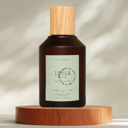 A brown glass bottle of ISULA parfums Filletta perl'Omu - Fern for Men eau de toilette with a wooden cap sits on a round wood platform, its light green label reading ISULA parfums, softly lit by natural sunlight with blurred shadows behind.