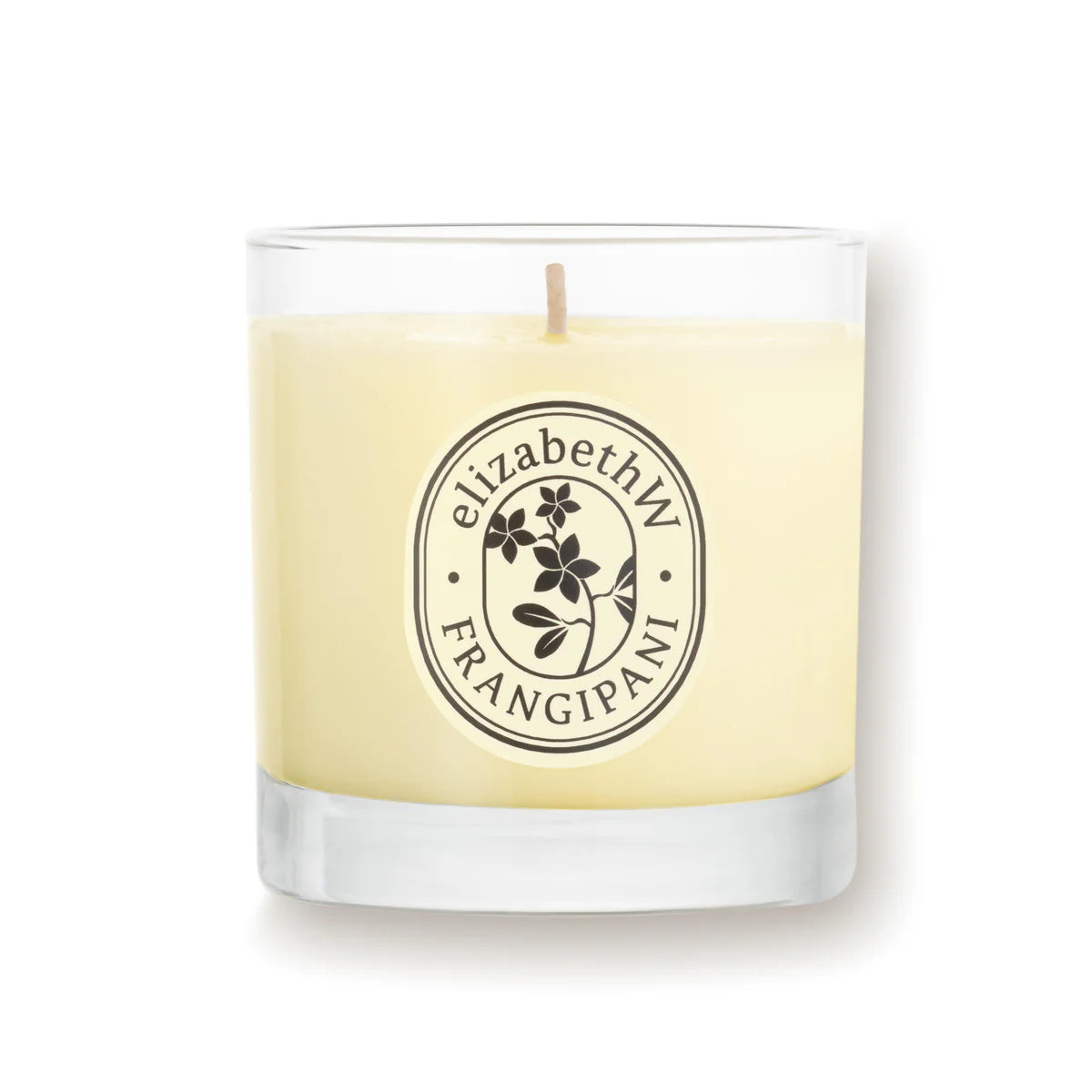 A glass jar candle from elizabeth W's Botanist's Scent Lab collection, labeled "Frangipani Candle," features a floral emblem. It is crafted with creamy soy wax and has a central wick, set against a white background.