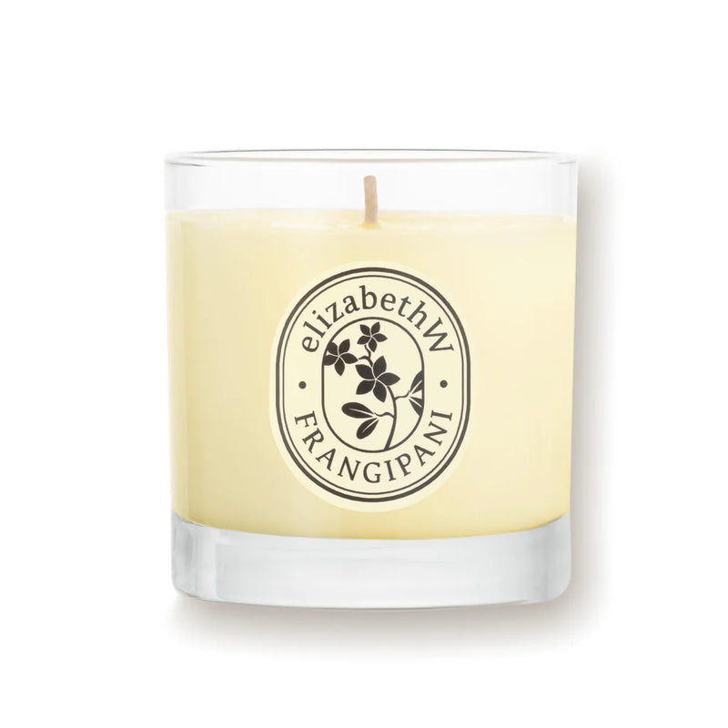 A glass jar candle from elizabeth W's Botanist's Scent Lab collection, labeled "Frangipani Candle," features a floral emblem. It is crafted with creamy soy wax and has a central wick, set against a white background.