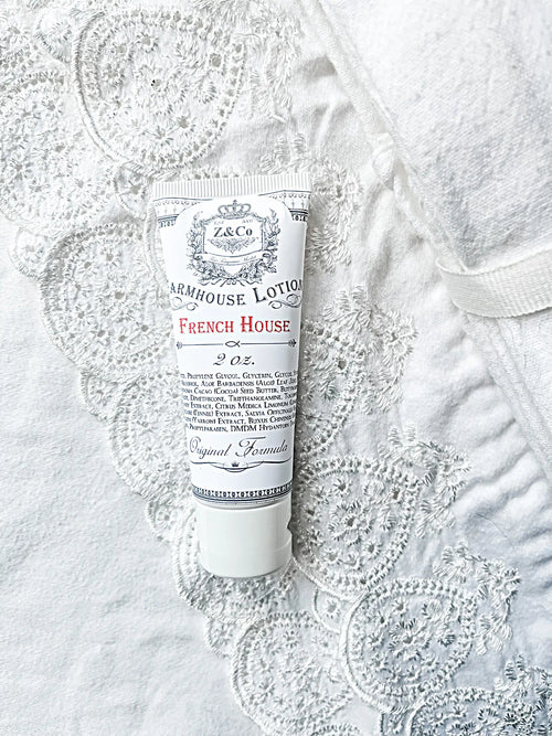 A tube of Z&Co. French House Farmhouse Lotion (2 oz.) rests gracefully on an intricately patterned lacy white fabric. The vintage-style design of the tube, featuring detailed ingredient information and the product's original formula, makes it perfect for on-the-go moisture needs. Both the tube and the cap are white, harmonizing elegantly with the fabric beneath it.