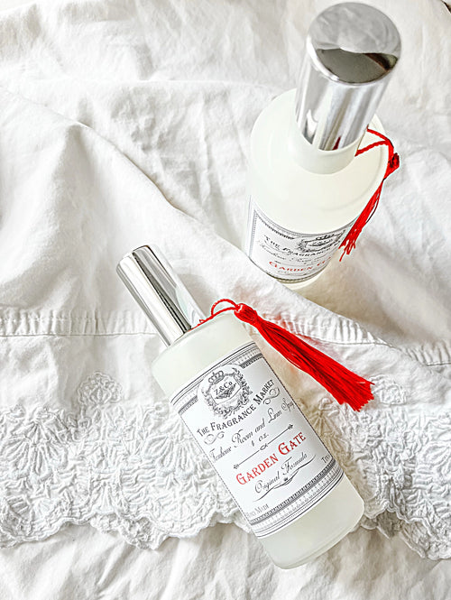 Two elegant Z&Co. Garden Gate Farmhouse Room/Linen Spray bottles rest on white textured fabric. With sophisticated labels, silver caps, and red tassels, the Gardenia and Tuberose fragrances evoke luxury. The intricate "Garden Gate" label ensures each is perfect as a room/linen spray choice.