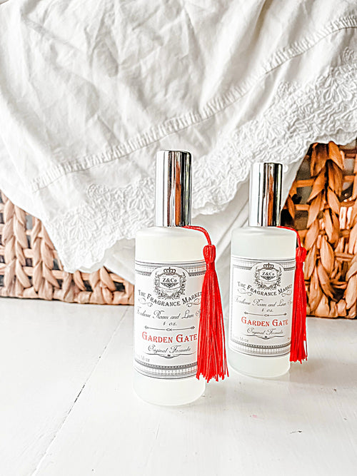 Two bottles of Z&Co. Garden Gate Farmhouse Room/Linen Spray rest on a white surface, each infused with gardenia and tuberose, featuring a silver cap and red tassel. A woven basket and white fabric softly blur in the background, enhancing the elegance.