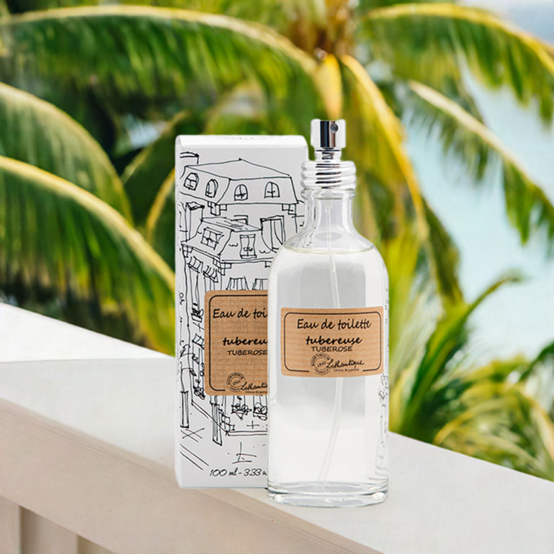 A bottle of Lothantique Tuberose Eau de Toilette stands on a white railing, next to its illustrated box. Palm fronds are visible in the blurred background, suggesting a tropical setting. This modern fragrance by Lothantique captures the timeless spirit of France with every spritz.