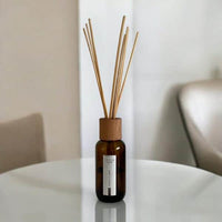 The ISULA parfums Room Diffuser - Dolce Farniente No.28 features a brown glass bottle with a wooden cap and reeds, highlighting artisanal craftsmanship and a woody-oriental scent, set on a round white table with blurred chairs in the background.
