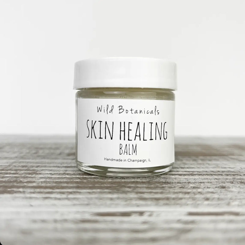 A jar of "Wild Botanicals Skin Healing Balm 1oz Glass Jar" by Wild Botanicals on a wooden surface with a white cap and clear label, handcrafted in Champaign, IL.