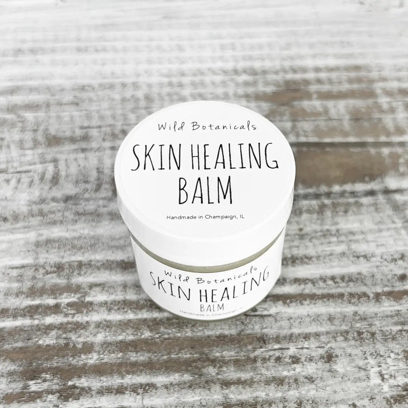 A small tin of "Wild Botanicals Skin Healing Balm 1oz Glass Jar" on a rustic wooden surface. The label states it's handmade in Champaign, IL.