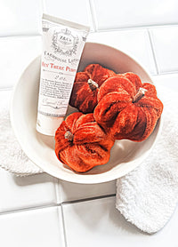 A white bowl containing three small, velvet-textured orange pumpkins and a travel-sized tube of Z&Co Hey There Pumpkin Farmhouse Lotion. The bowl rests on a white countertop with a folded white towel partially tucked underneath.